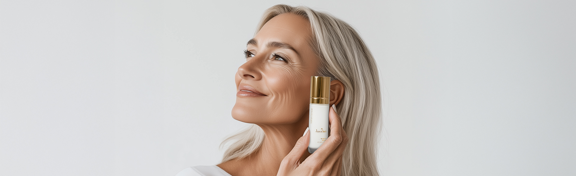Ältere Frau mit langen, grauen Haaren hält eine JEAN D'ARCEL Serum-Flasche und lächelt zur Seite.